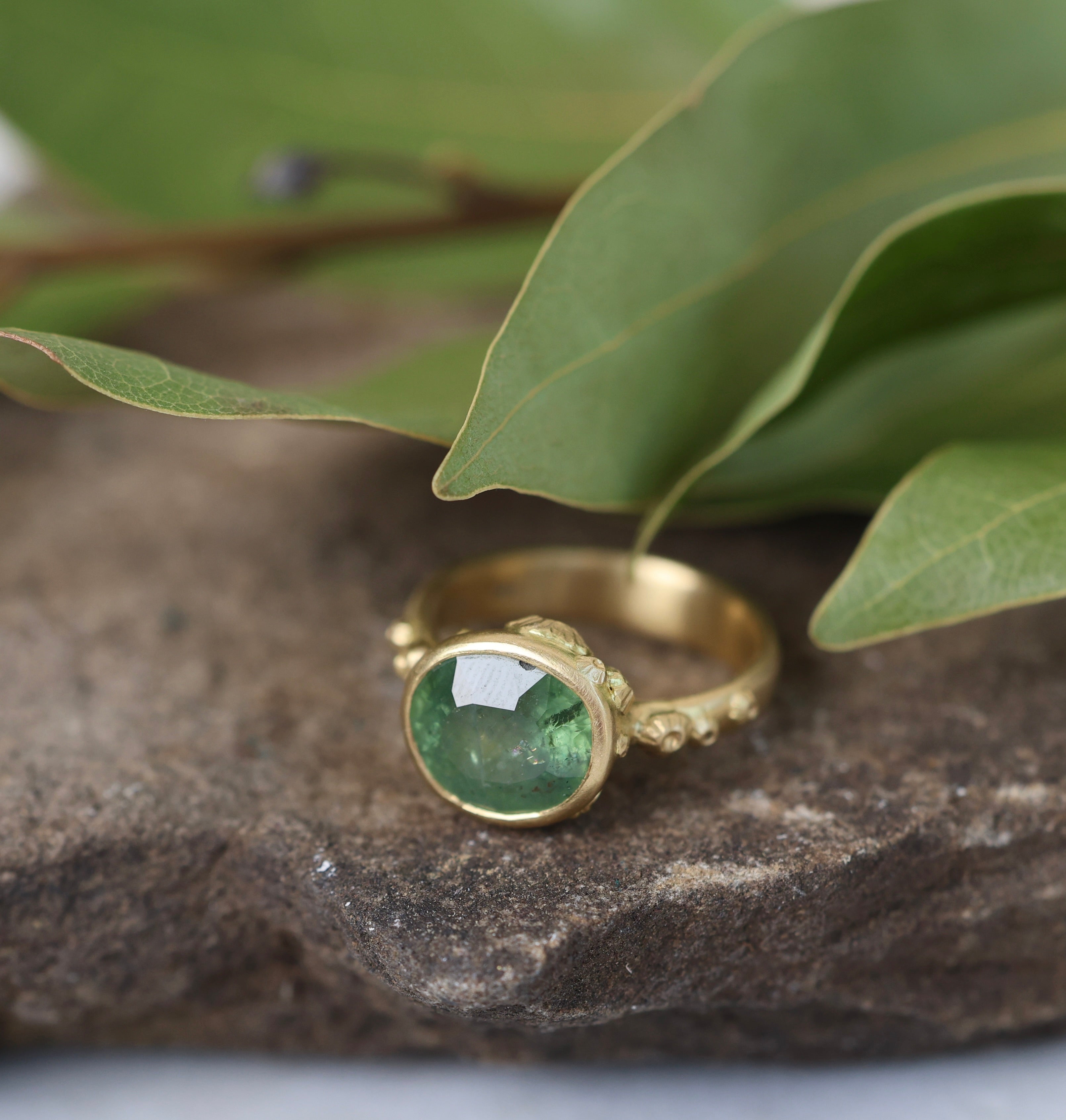Gold ring with a green gemstone on a stone surface with leaves in the background
