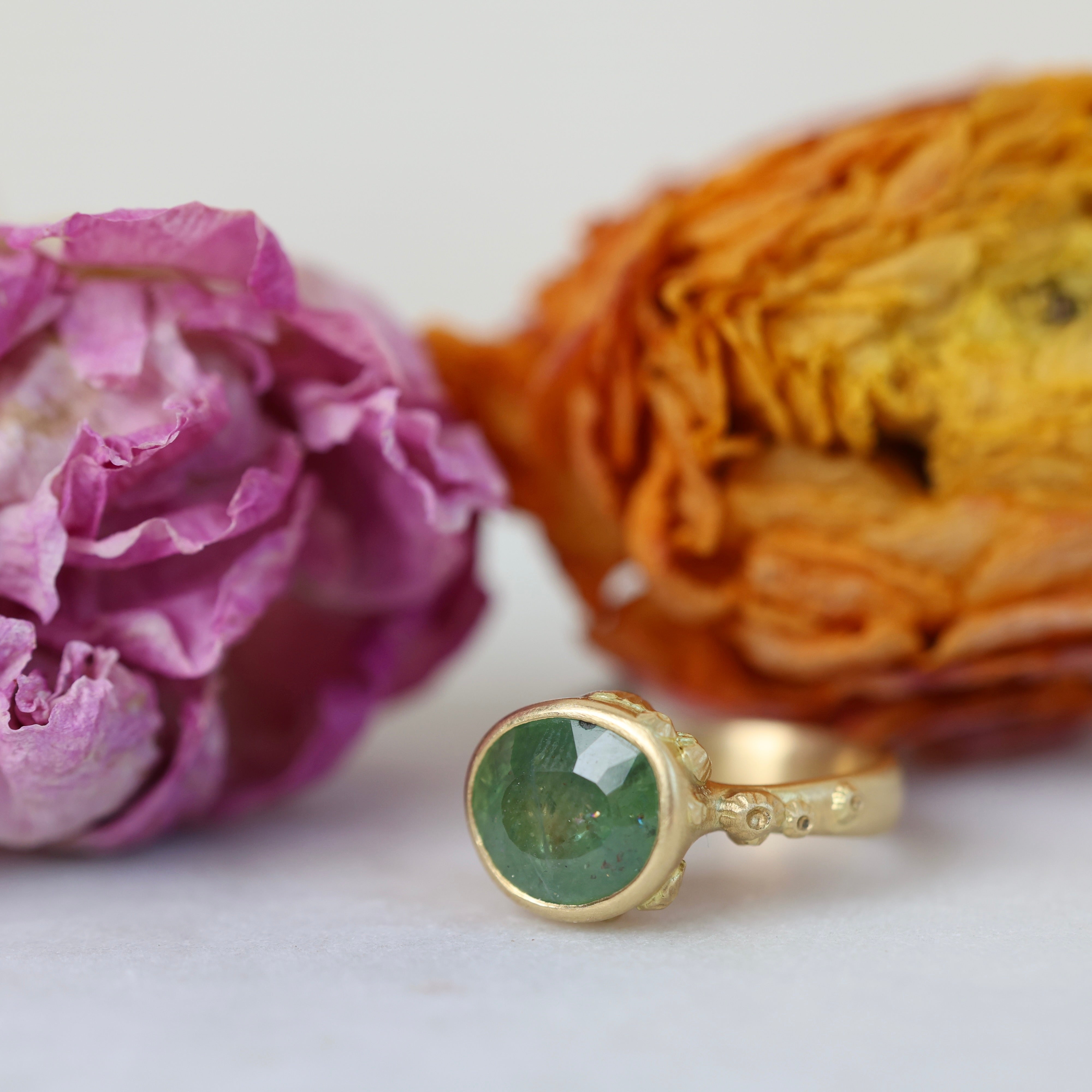 Gold ring with a green gemstone next to dried flowers on a white background