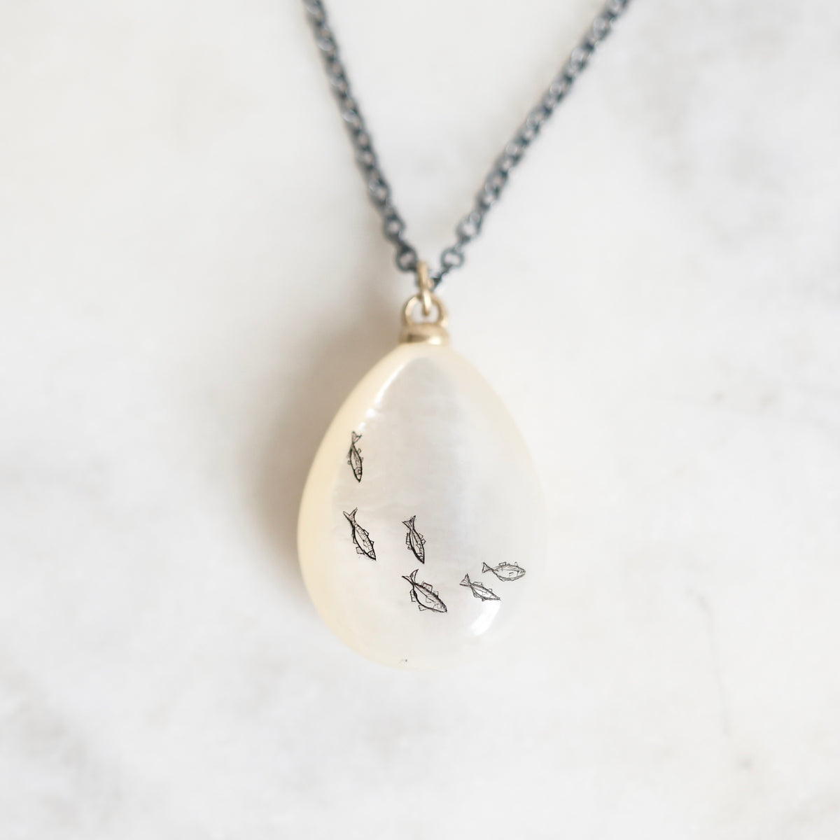 A teardrop-shaped pendant necklace made of milky white resin with small black fish outlines, hanging from a dark gray chain on a light marble surface.