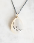 A teardrop-shaped pendant necklace made of milky white resin with small black fish outlines, hanging from a dark gray chain on a light marble surface.
