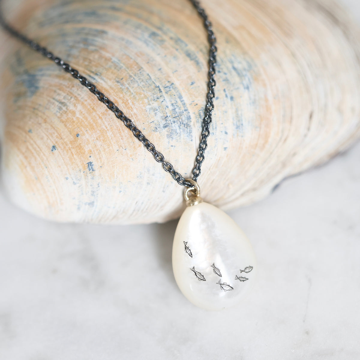 A close-up of a necklace with a dark chain and a teardrop-shaped white pendant featuring small fish designs, resting on a seashell over a light marble surface.