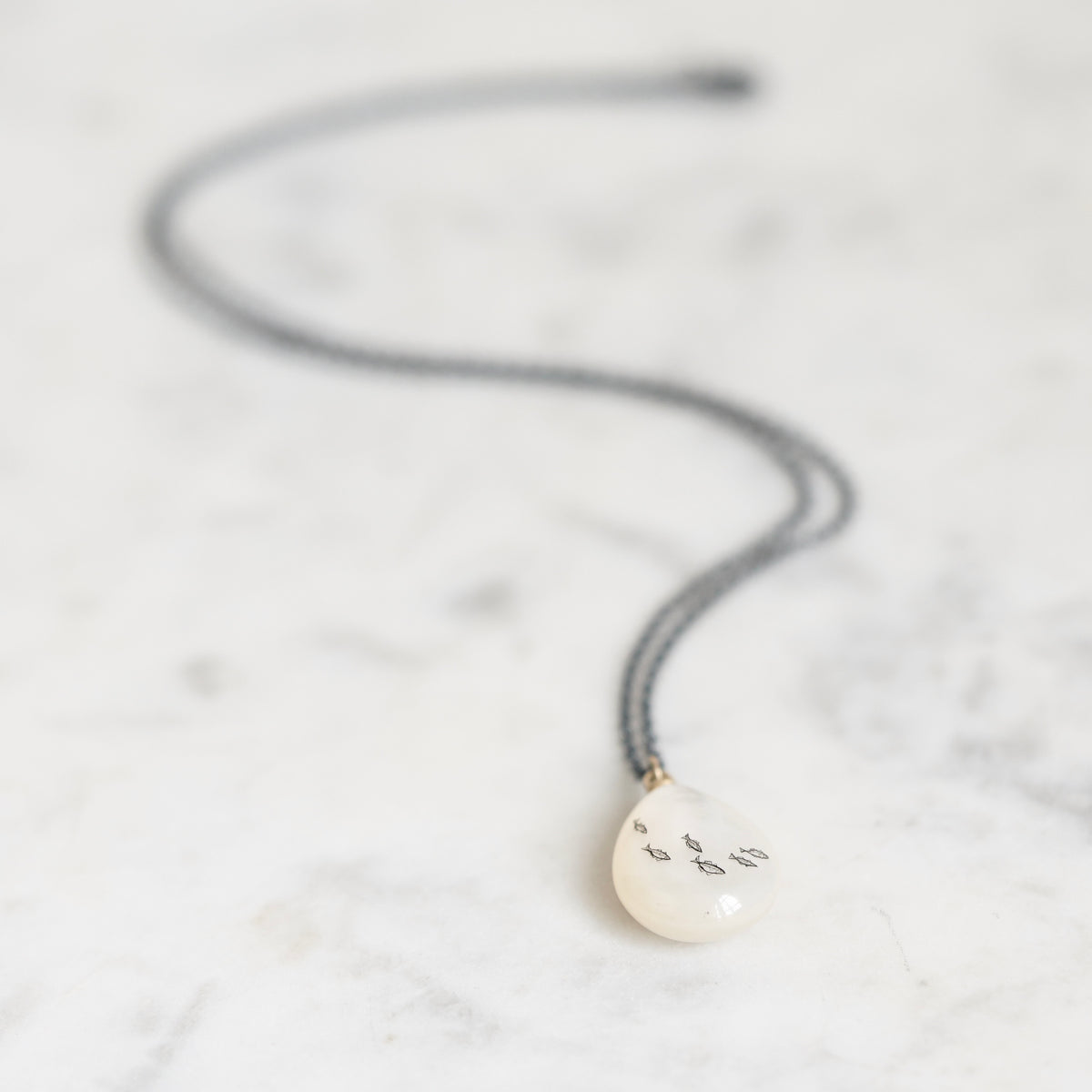 A teardrop-shaped white pendant necklace with hand-painted black symbols, hanging from a dark gray cord on a light marble surface.