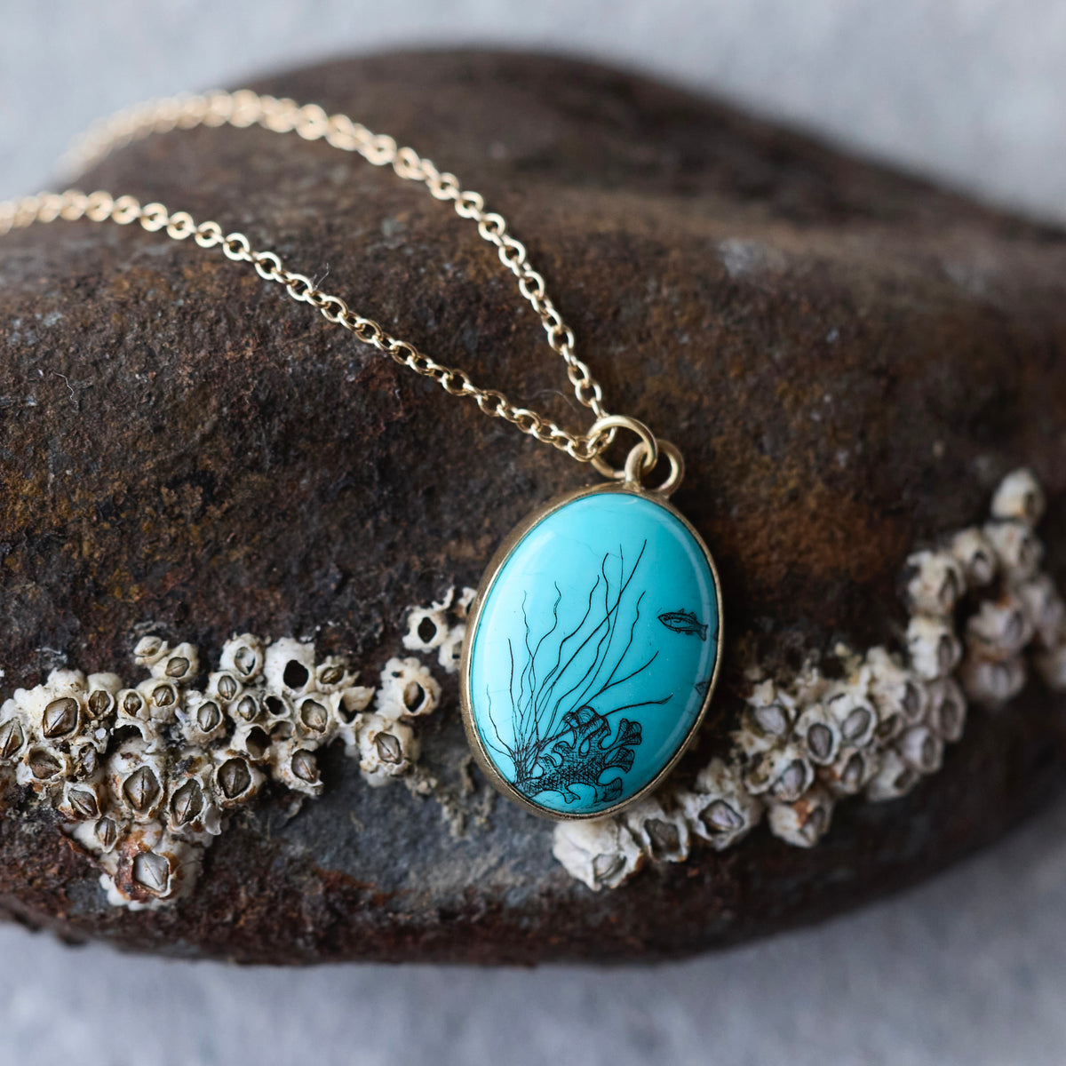 Gold chain necklace with an oval turquoise pendant featuring black fish and coral designs, displayed on a dark rock with sea urchin shells.