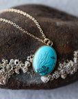 Gold chain necklace with an oval turquoise pendant featuring black fish and coral designs, displayed on a dark rock with sea urchin shells.