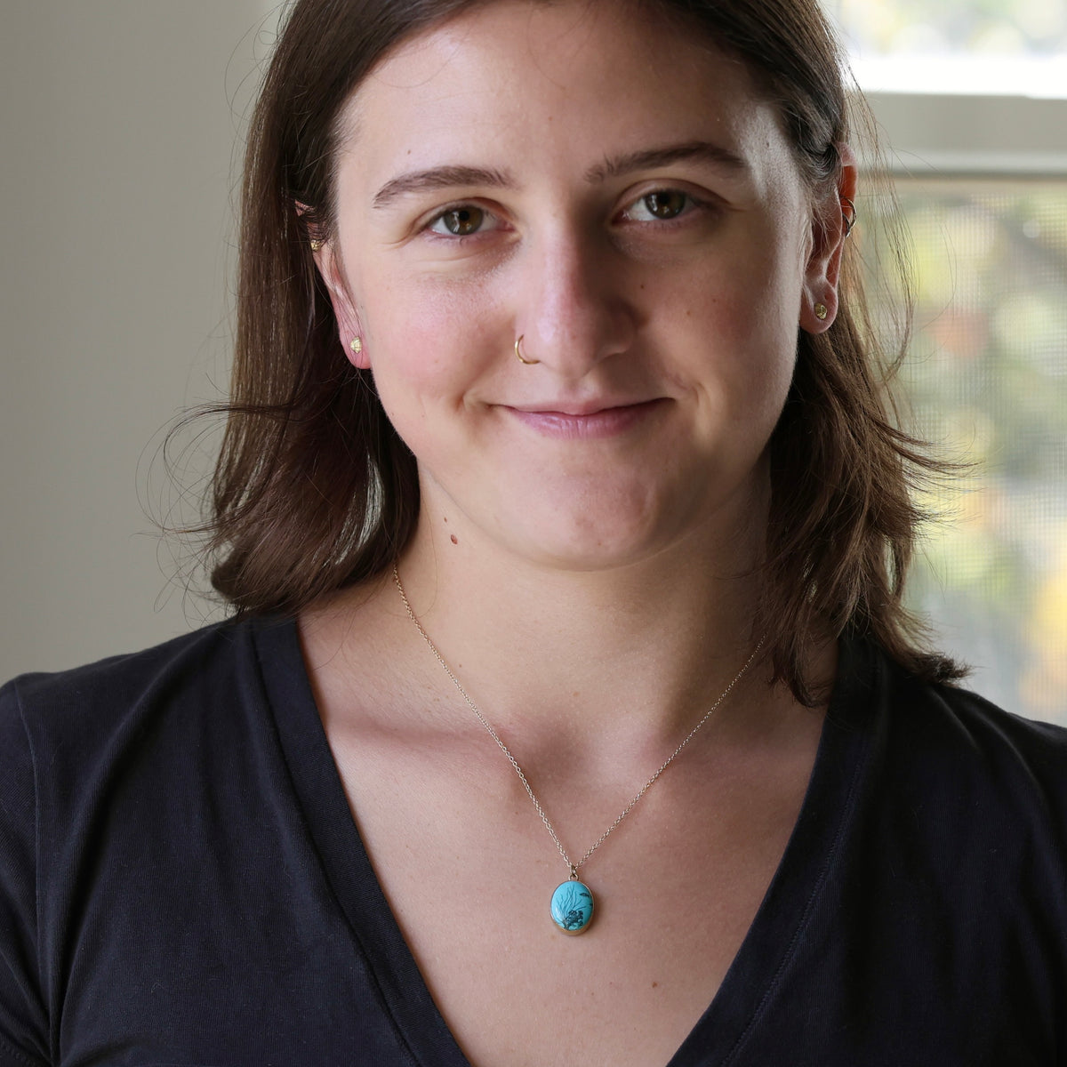 A woman with brown hair wearing a silver chain necklace with a round turquoise pendant, photographed in a close-up portrait against a softly blurred indoor background.