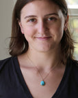 A woman with brown hair wearing a silver chain necklace with a round turquoise pendant, photographed in a close-up portrait against a softly blurred indoor background.