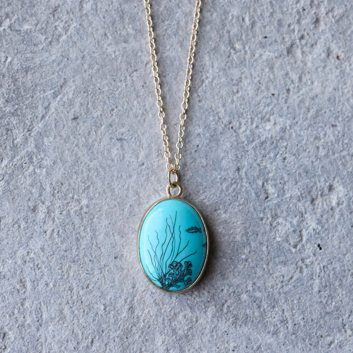 An oval pendant necklace featuring a turquoise blue background with black underwater coral and fish designs, suspended from a delicate gold chain against a textured gray background.