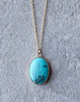 An oval pendant necklace featuring a turquoise blue background with black underwater coral and fish designs, suspended from a delicate gold chain against a textured gray background.