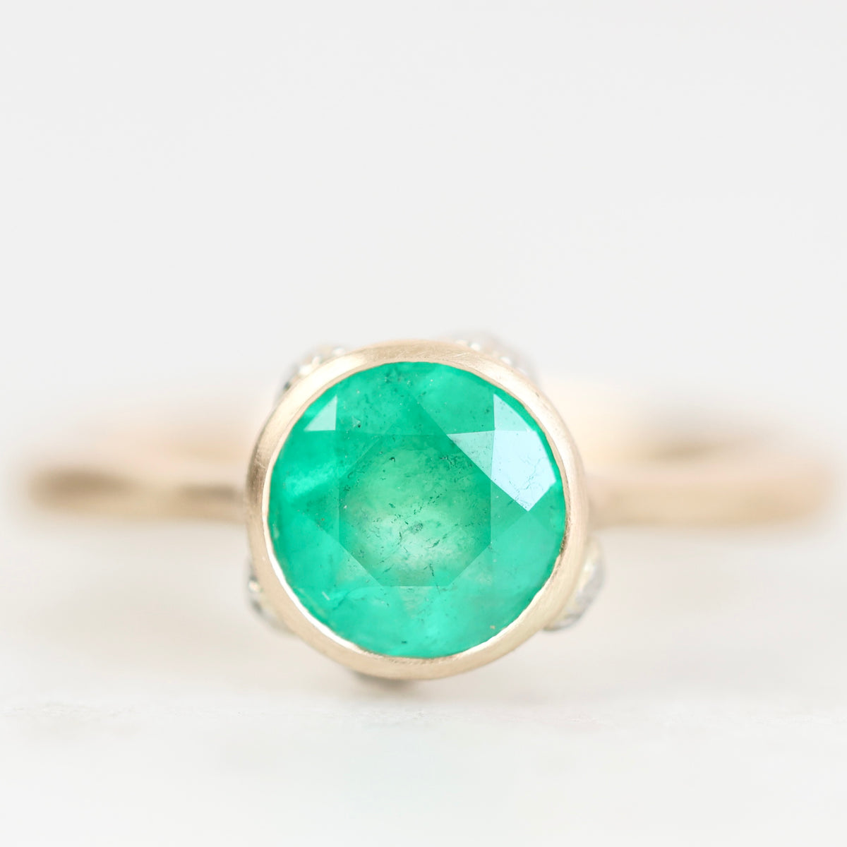 A round-cut emerald green gemstone ring set in a gold bezel, shown on a clean white background with a soft shallow depth of field.