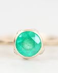 A round-cut emerald green gemstone ring set in a gold bezel, shown on a clean white background with a soft shallow depth of field.