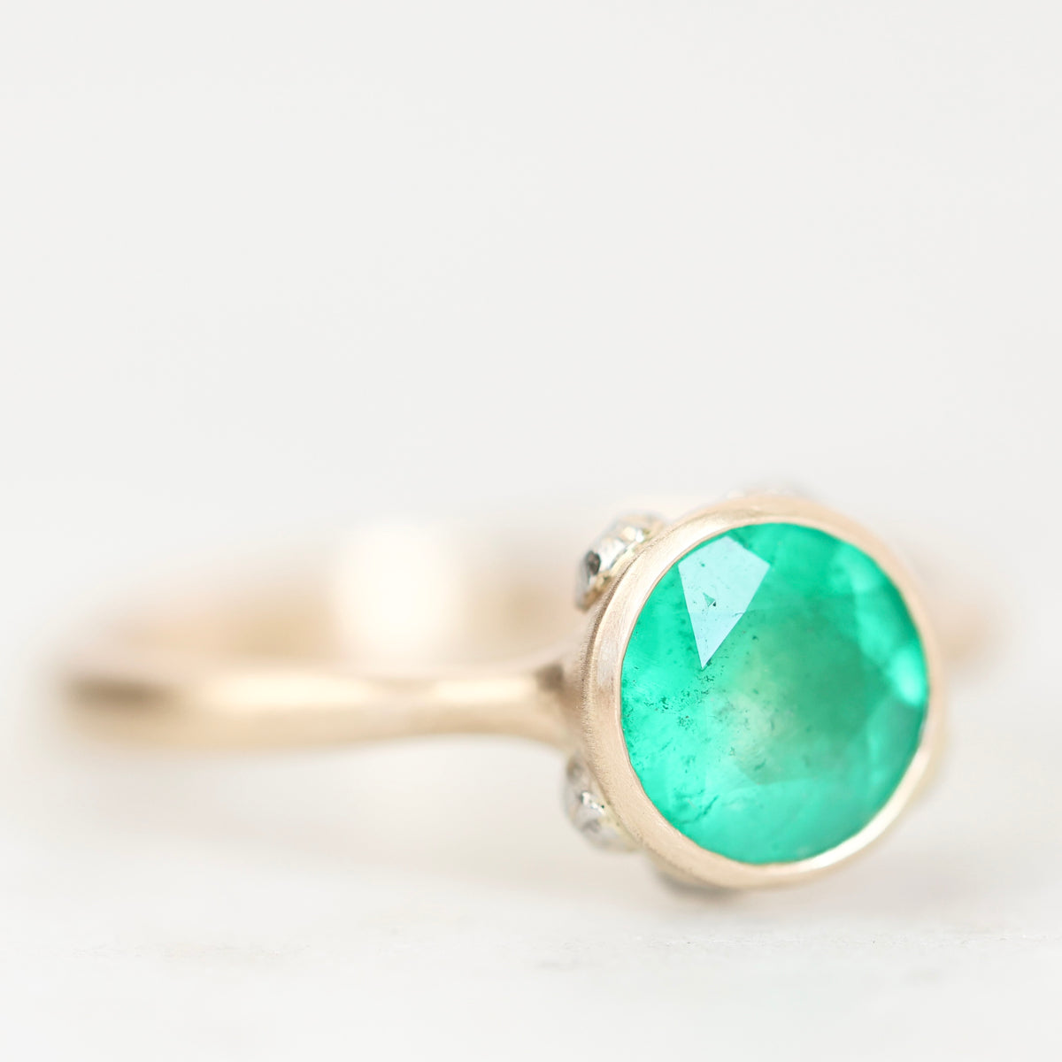 A gold ring featuring a round, vibrant green emerald-cut gemstone, accented by small clear stones on the band, set against a soft white background.