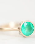 A gold ring featuring a round, vibrant green emerald-cut gemstone, accented by small clear stones on the band, set against a soft white background.