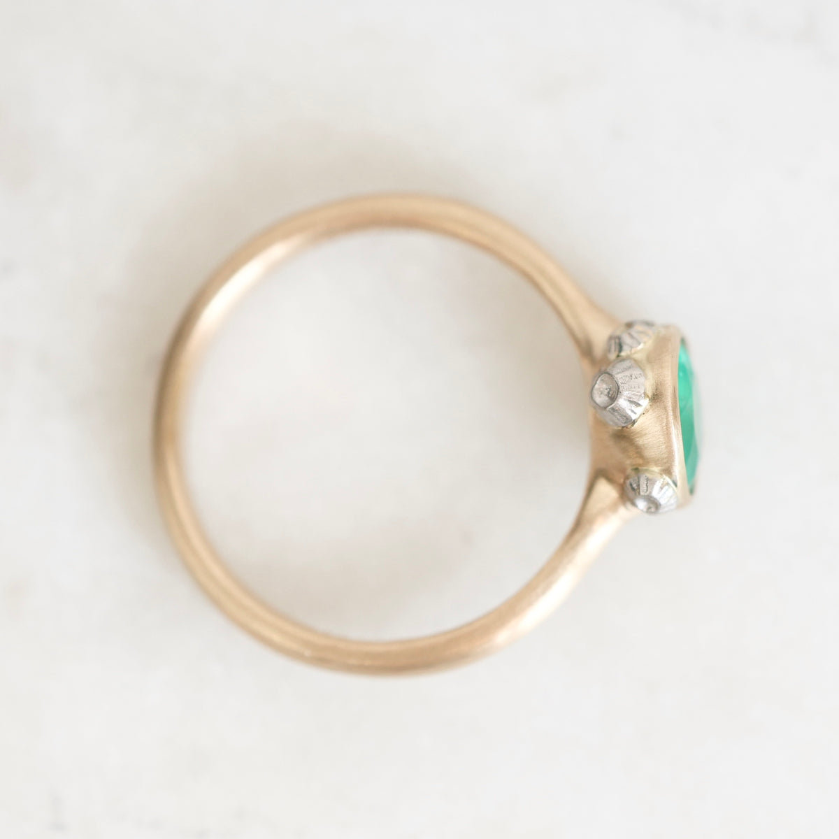 A gold ring with a green oval gemstone and small clear accent stones, photographed from above on a light marble-like surface.