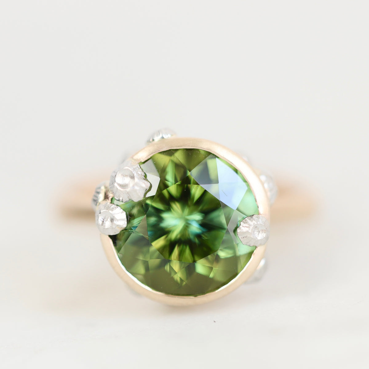 A round-cut green gemstone ring set in a gold-toned bezel with small clear accent stones, photographed on a clean light gray background.
