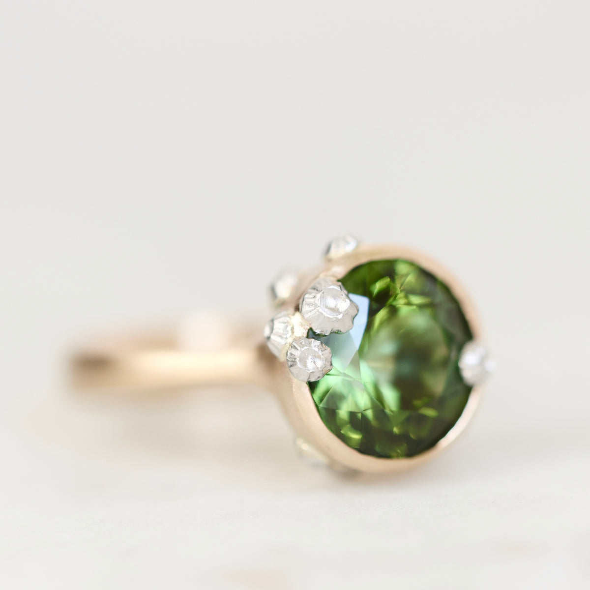 A close-up shot of a ring featuring a round green gemstone in a gold-toned setting, accented with small clear stones, on a light gray background.