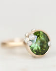 A close-up shot of a ring featuring a round green gemstone in a gold-toned setting, accented with small clear stones, on a light gray background.