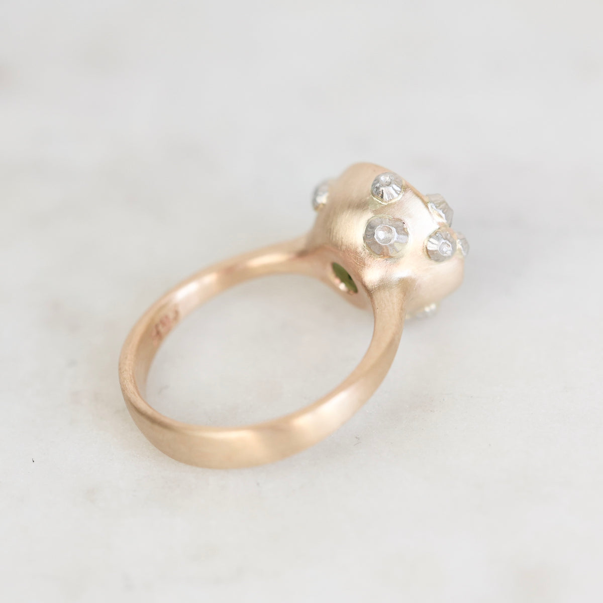 A close-up shot of a rose gold ring with a sculptural flower head setting adorned with small clear gemstones, resting on a light marble surface.
