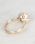 A close-up shot of a rose gold ring with a sculptural flower head setting adorned with small clear gemstones, resting on a light marble surface.