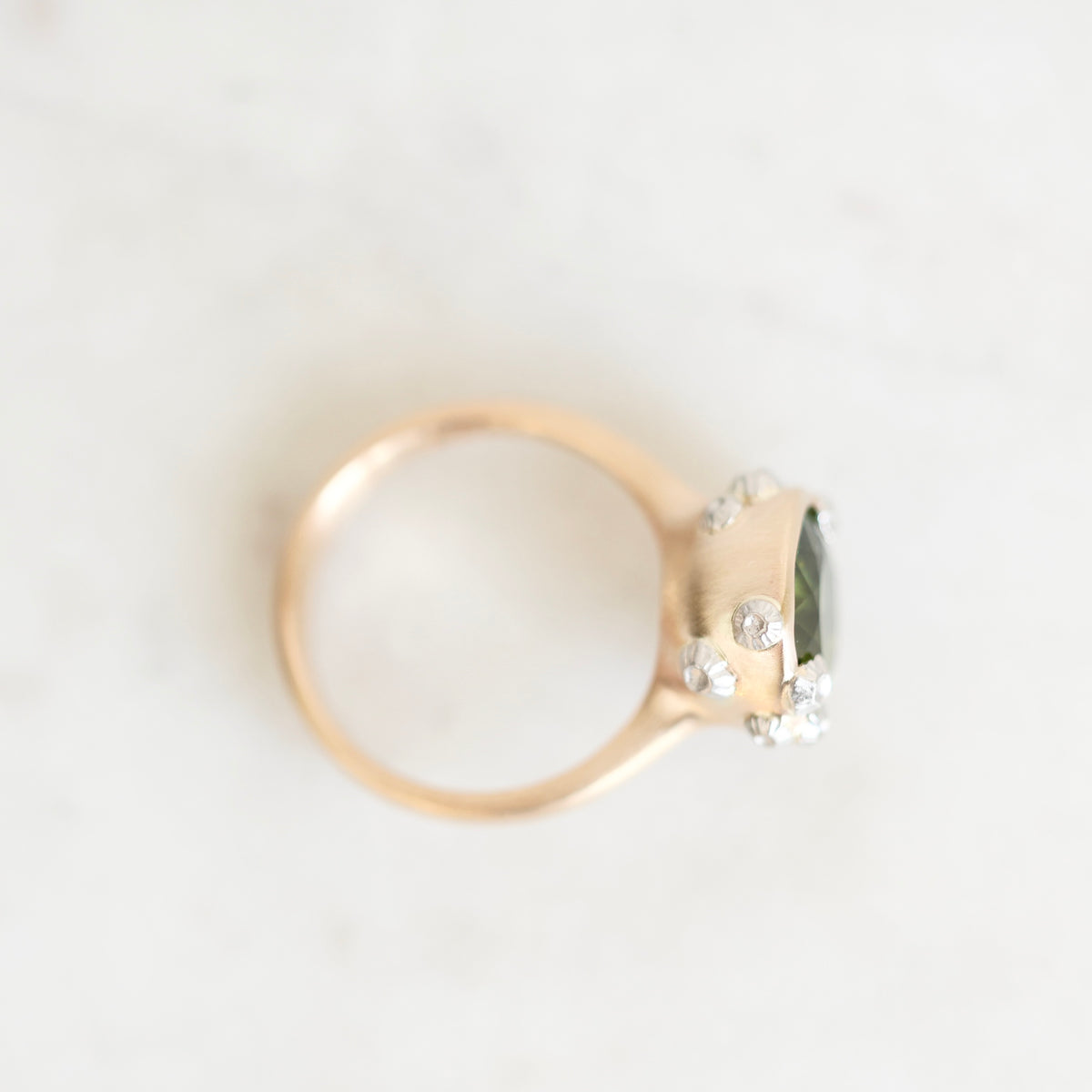 A gold ring with a cluster of clear stones and a dark green rectangular gemstone, photographed on a light marble surface.