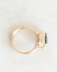 A gold ring with a cluster of clear stones and a dark green rectangular gemstone, photographed on a light marble surface.