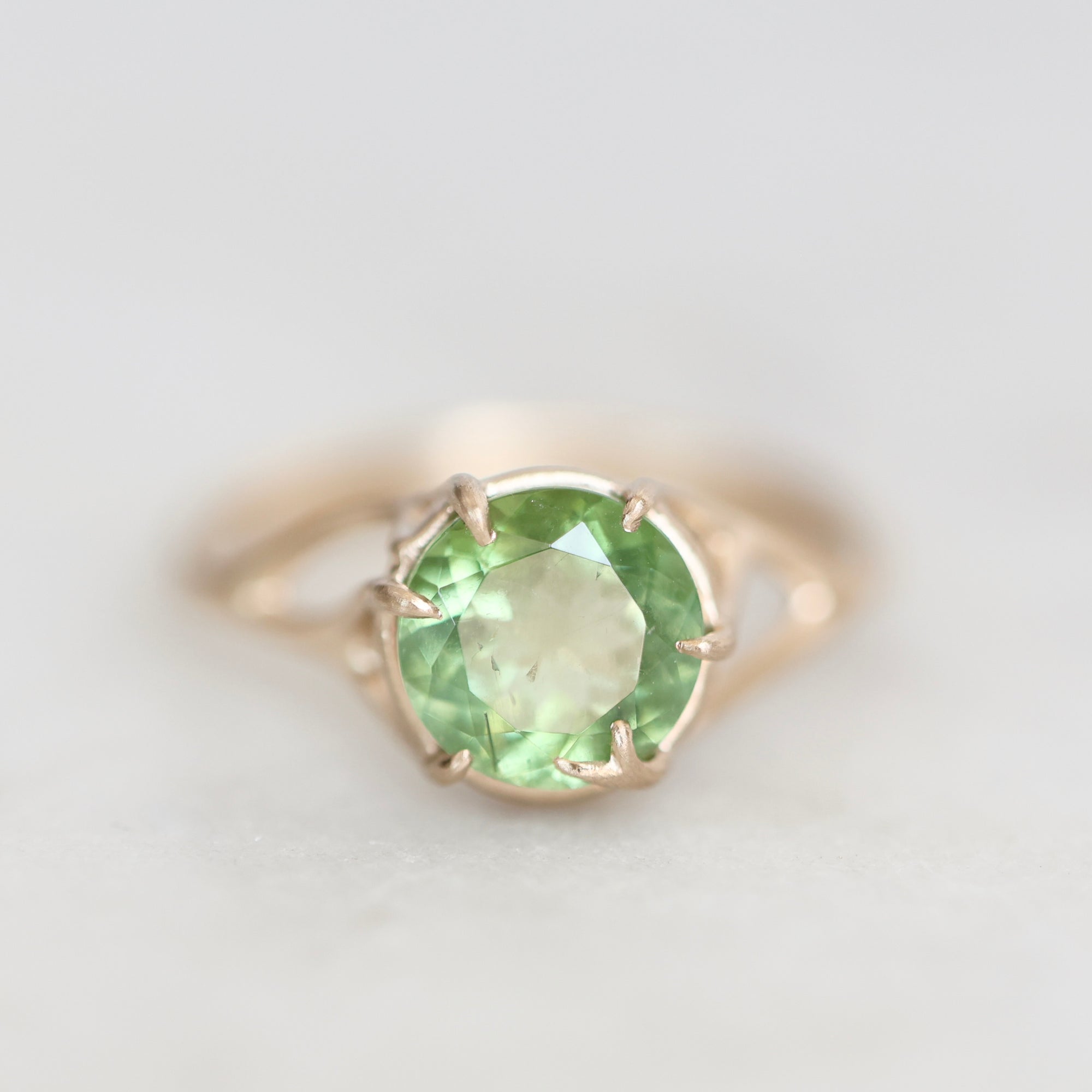 A round-cut light green gemstone ring set in a gold prong setting, shown on a soft light gray background.
