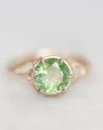 A round-cut light green gemstone ring set in a gold prong setting, shown on a soft light gray background.