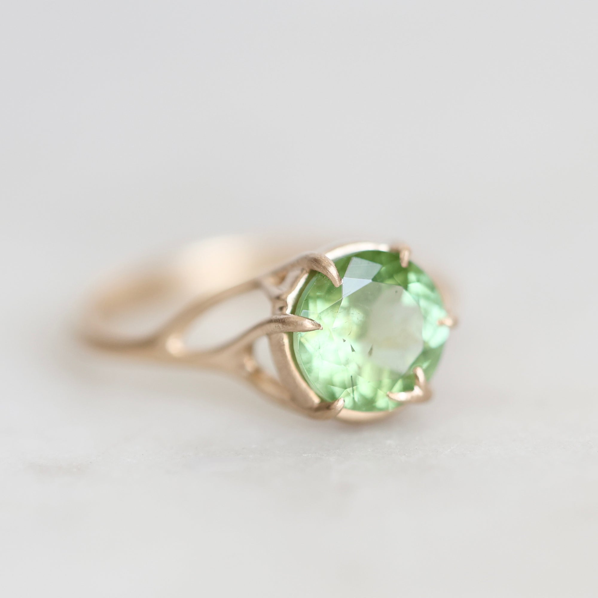A gold ring featuring a round-cut light green gemstone in a prong setting, shown on a soft, light gray background.