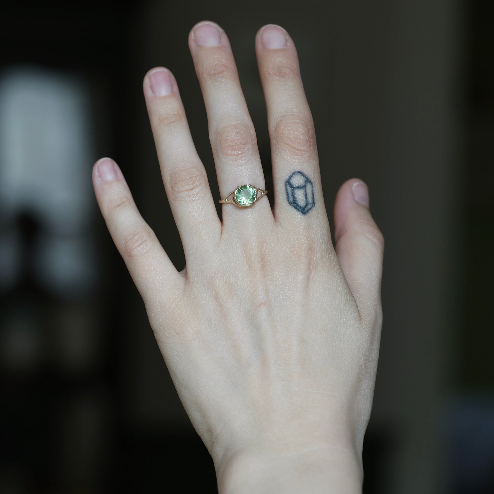 A close-up of a person's hand wearing a gold ring with a green oval gemstone, with a small black geometric tattoo on the side of the hand.