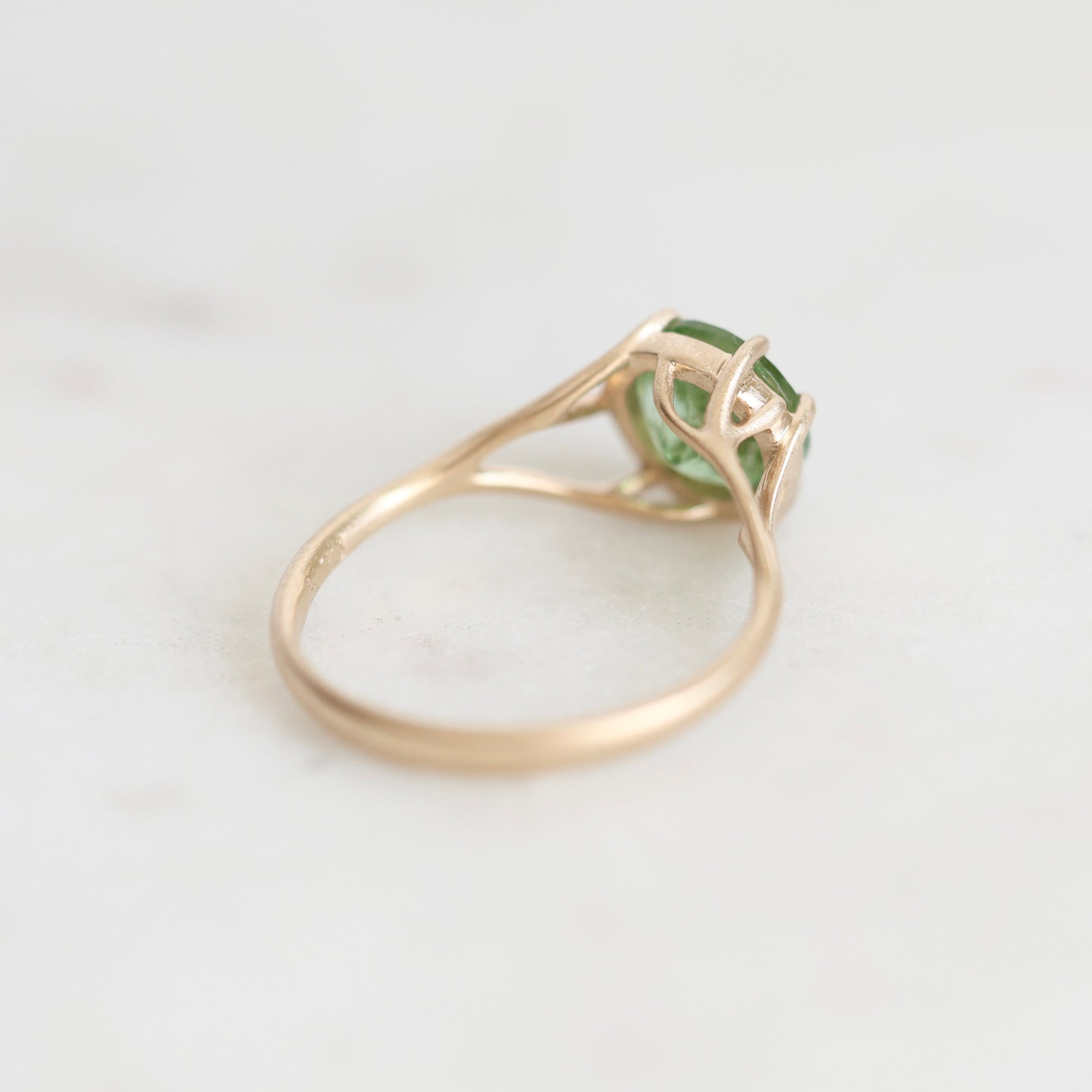 A gold ring with a green gemstone in a prong setting, photographed on a light marble surface.