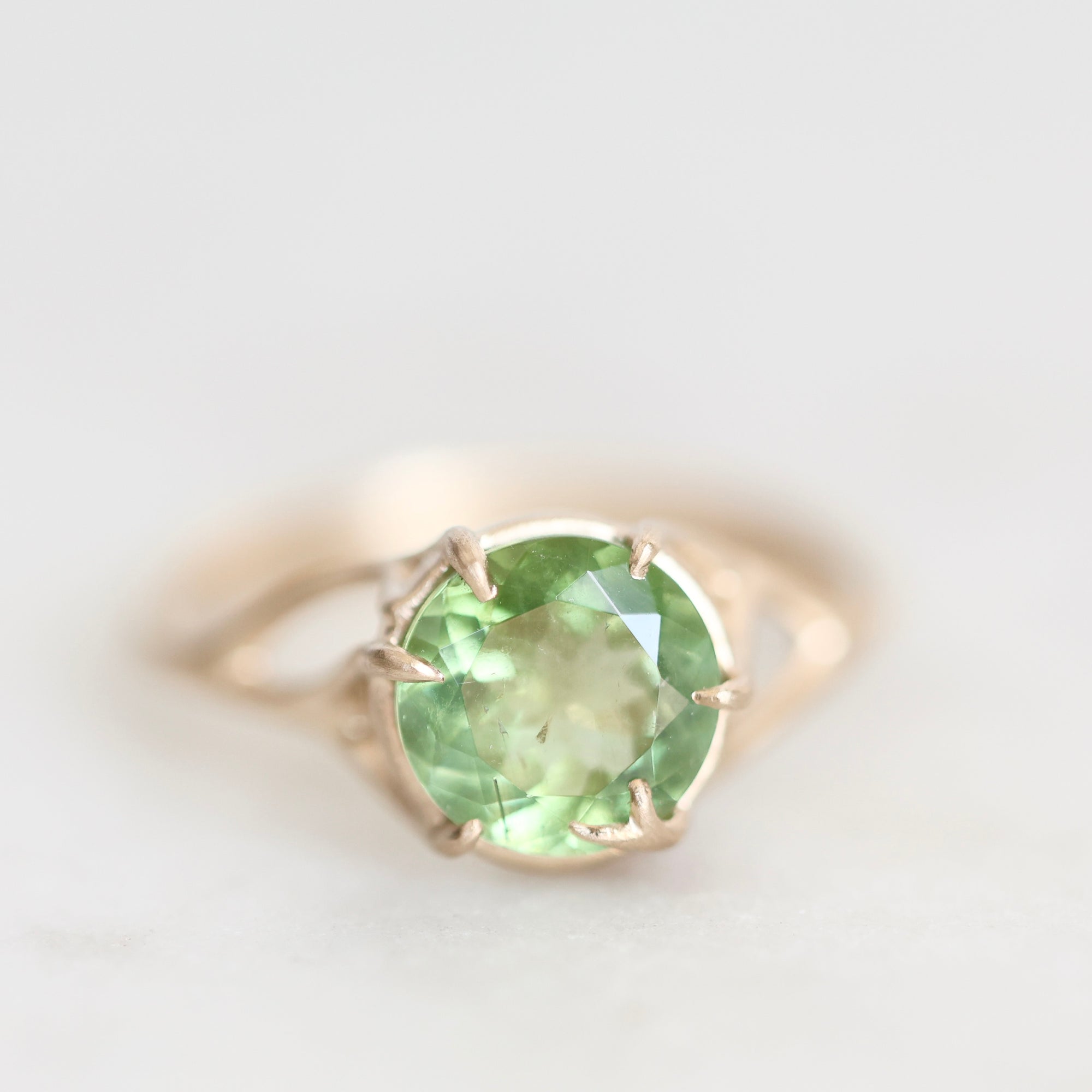 A round-cut light green gemstone ring set in a gold-toned prong setting on a soft white background.