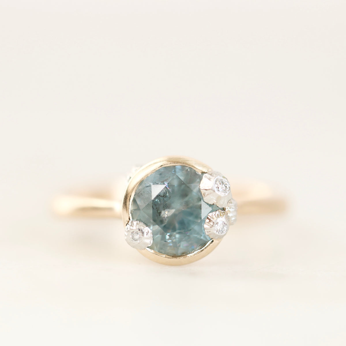 A gold ring featuring a round-cut light blue gemstone in a bezel setting, accented by three small clear stones on the right side, photographed against a soft white background.