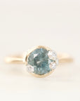 A gold ring featuring a round-cut light blue gemstone in a bezel setting, accented by three small clear stones on the right side, photographed against a soft white background.