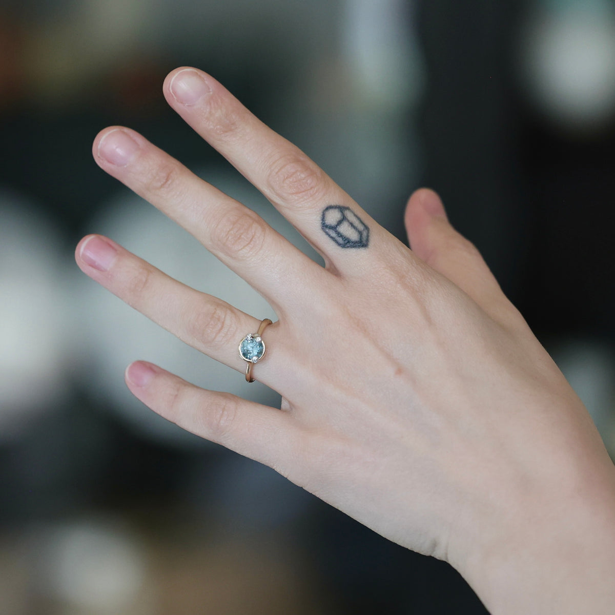 A close-up of a person's hand wearing a delicate gold ring with a small oval light blue gemstone. The hand also features a small geometric tattoo on the middle finger, set against a dark, blurred background.