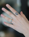 A close-up of a person's hand wearing a delicate gold ring with a small oval light blue gemstone. The hand also features a small geometric tattoo on the middle finger, set against a dark, blurred background.