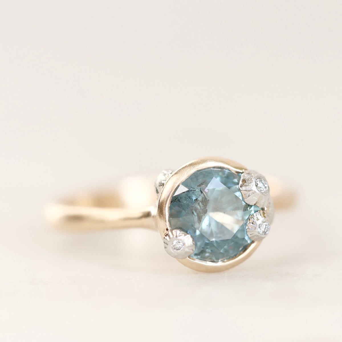 A gold ring featuring a round-cut light blue gemstone in a prong setting, accented with small clear stones on the shoulders, shown against a plain light background.