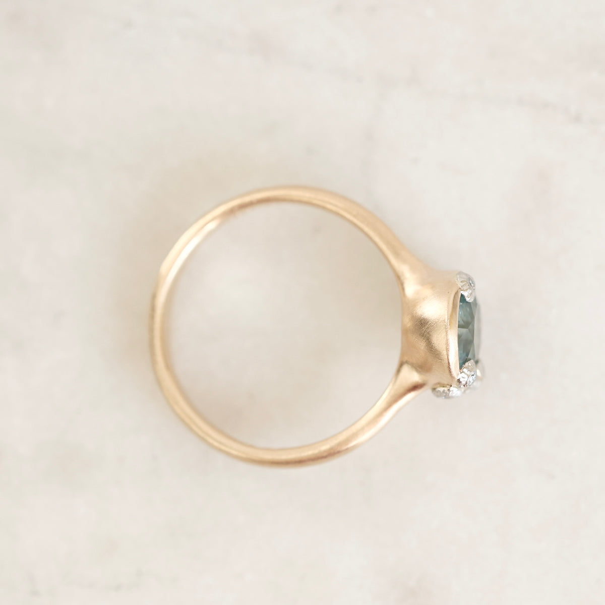A gold ring with a textured band and a bezel-set oval light blue gemstone accented by small clear stones, photographed on a light marble surface.