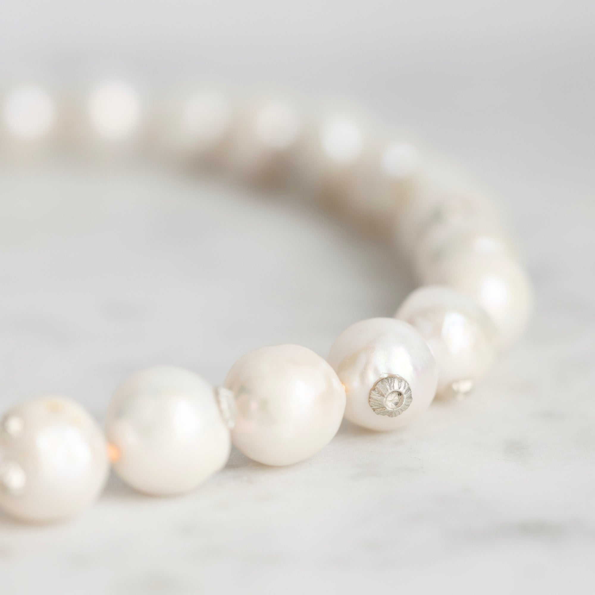 A close-up of a pearl beaded bracelet on a light marble surface, showing glossy white pearls and a small silver-toned spacer bead.