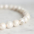 A close-up of a pearl beaded bracelet on a light marble surface, showing glossy white pearls and a small silver-toned spacer bead.