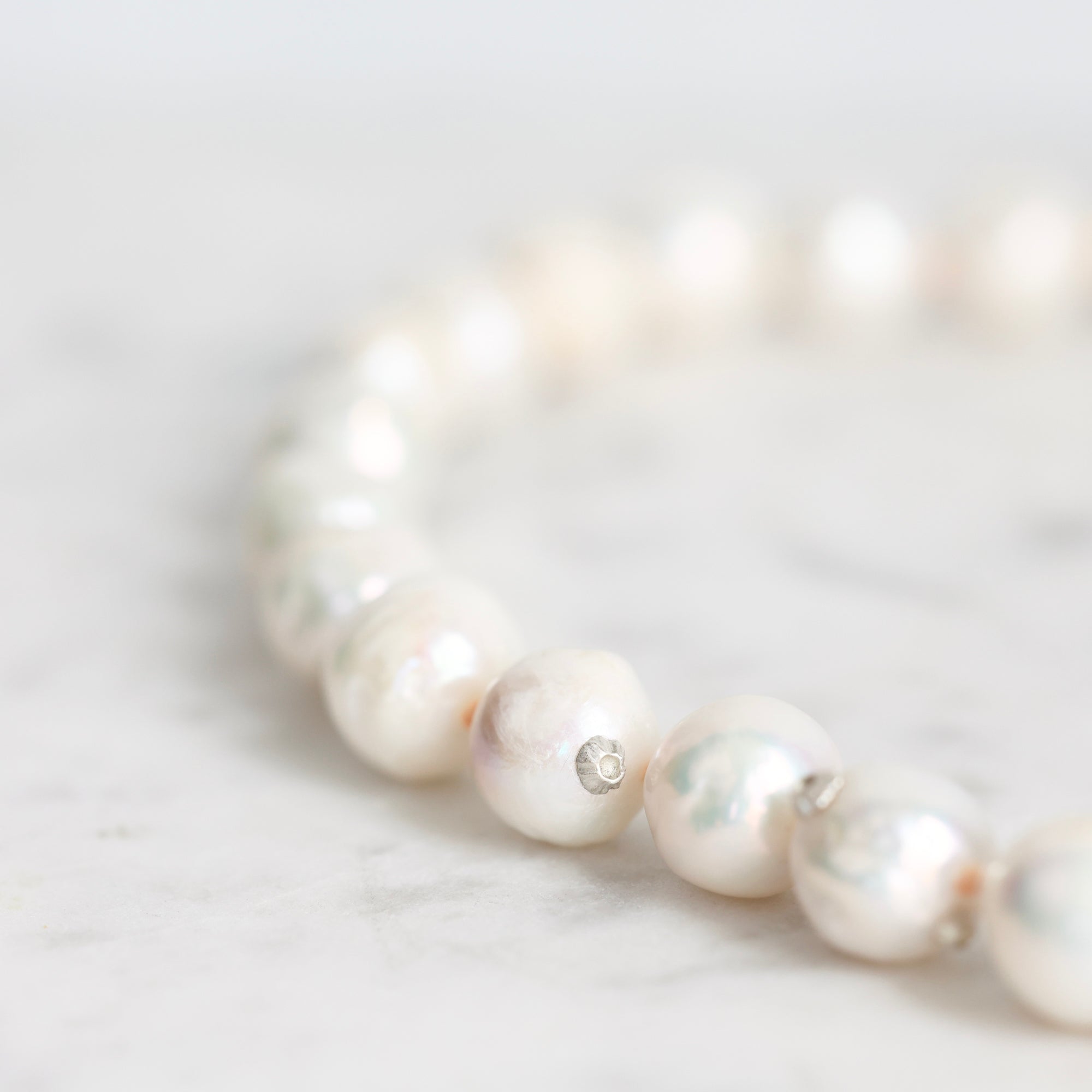 A close-up of a strand of irregular white baroque pearls on a light marble surface, with a small silver spacer bead visible.