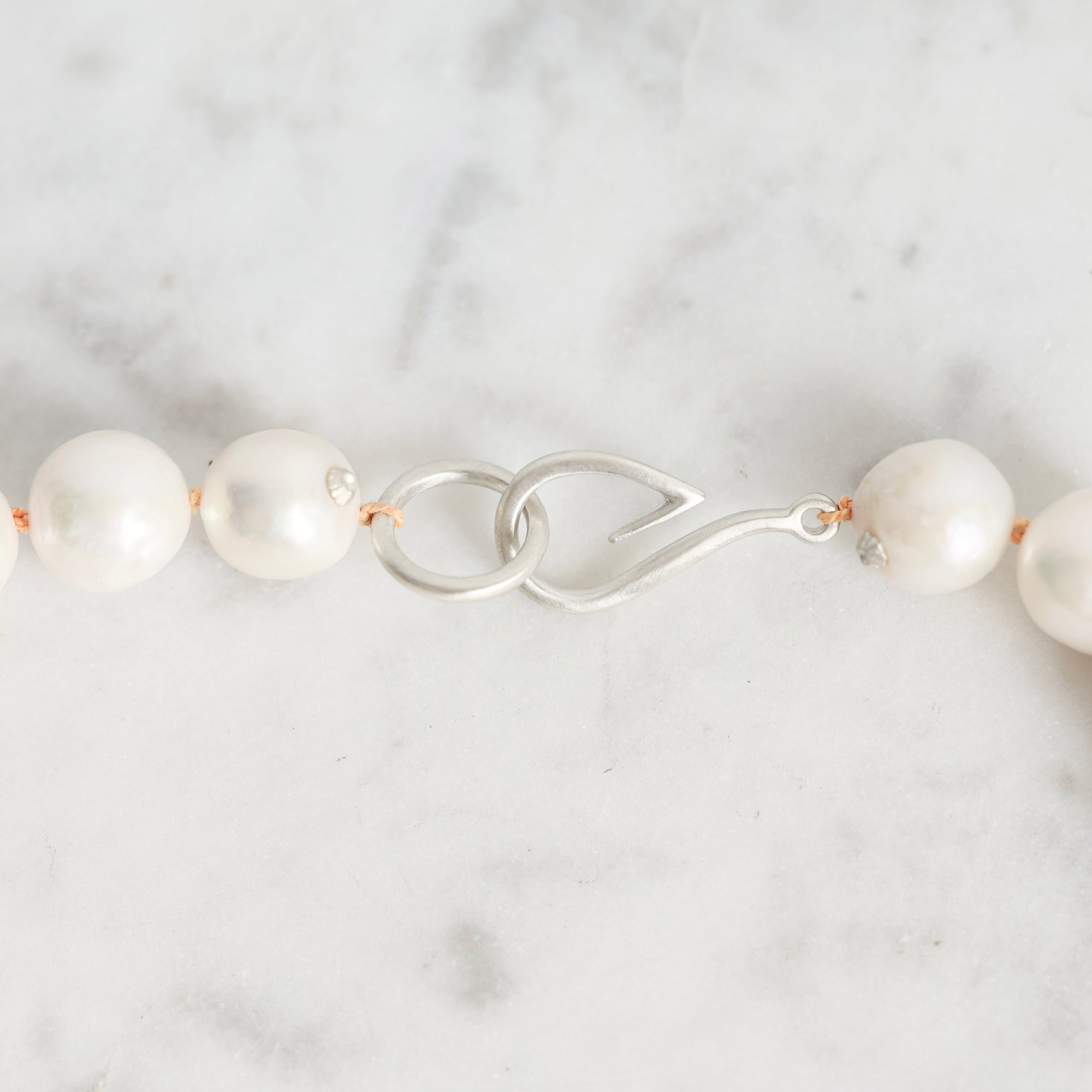 A close-up of a pearl necklace with a silver fishhook clasp on a light marble surface.