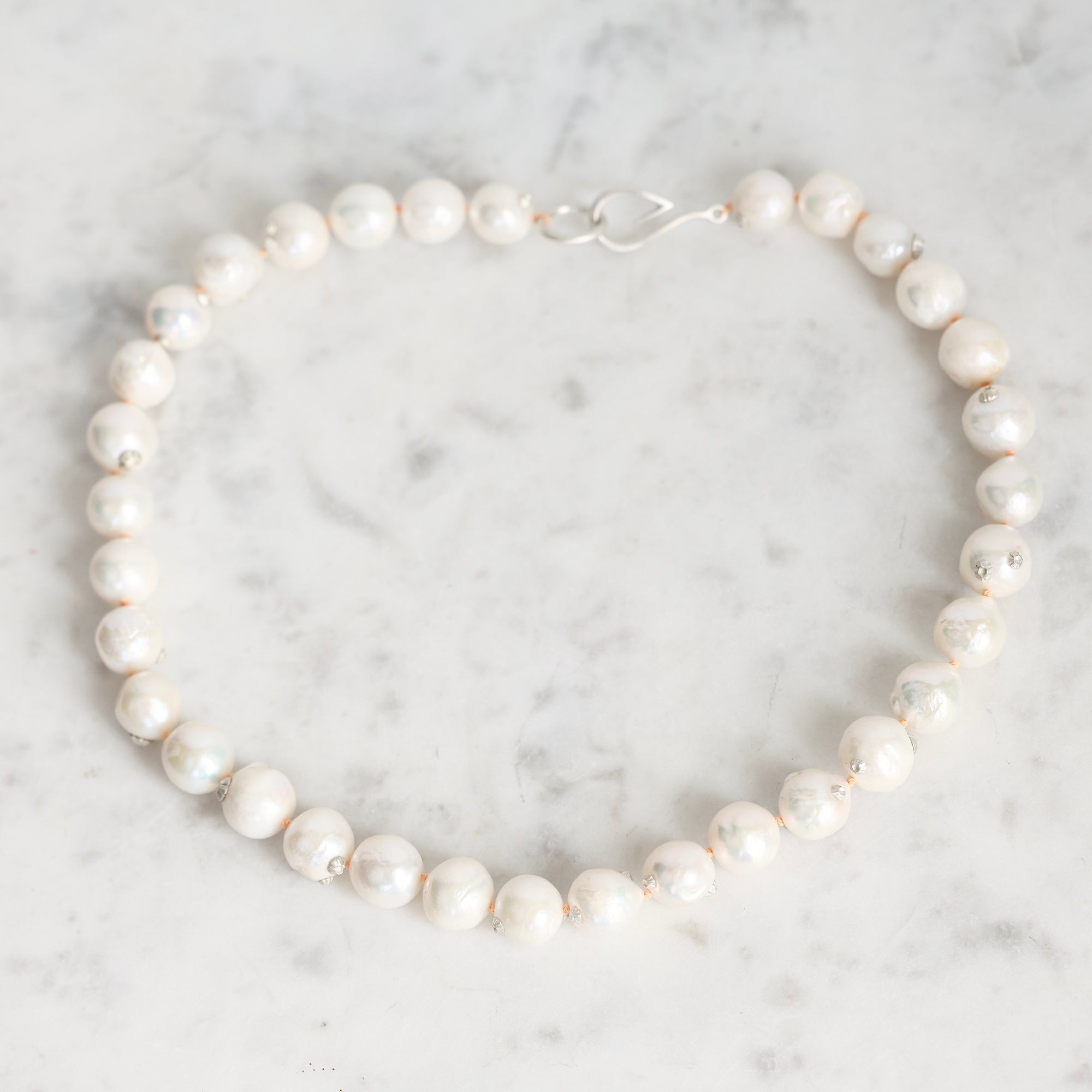 A strand of irregular white freshwater pearls with a silver clasp, arranged in a circle on a light gray and white marble surface.