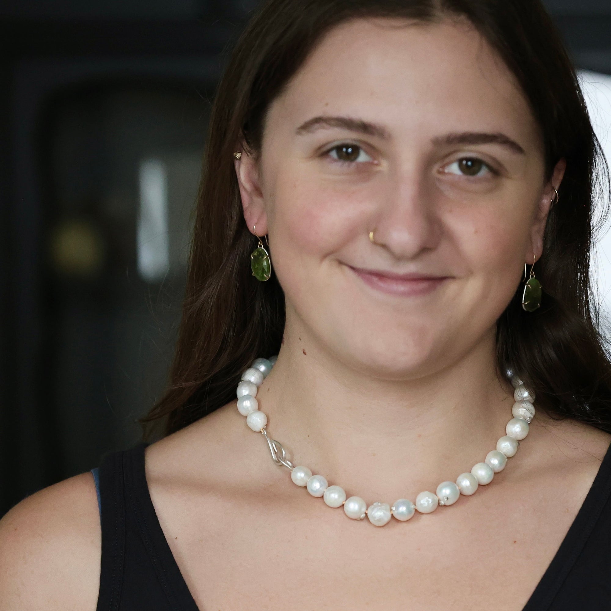 A close-up portrait of a woman wearing a pearl necklace with a silver clasp, matching green teardrop earrings, and a nose ring.