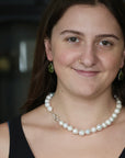 A close-up portrait of a woman wearing a pearl necklace with a silver clasp, matching green teardrop earrings, and a nose ring.