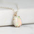 A close-up of a silver chain necklace with a round iridescent opal pendant in a gold-tone prong setting, resting on a white marble-like surface.