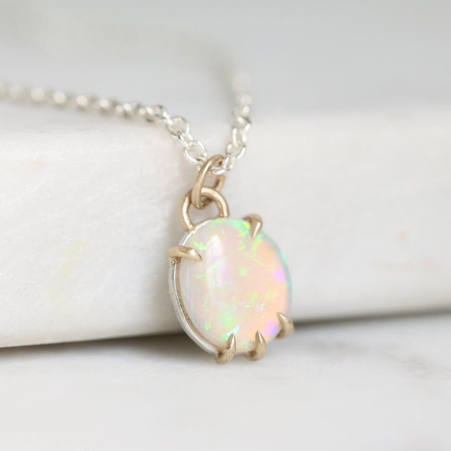 A close-up of a silver chain necklace with a round iridescent opal pendant in a gold-tone prong setting, resting on a white marble-like surface.