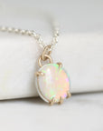A close-up of a silver chain necklace with a round iridescent opal pendant in a gold-tone prong setting, resting on a white marble-like surface.