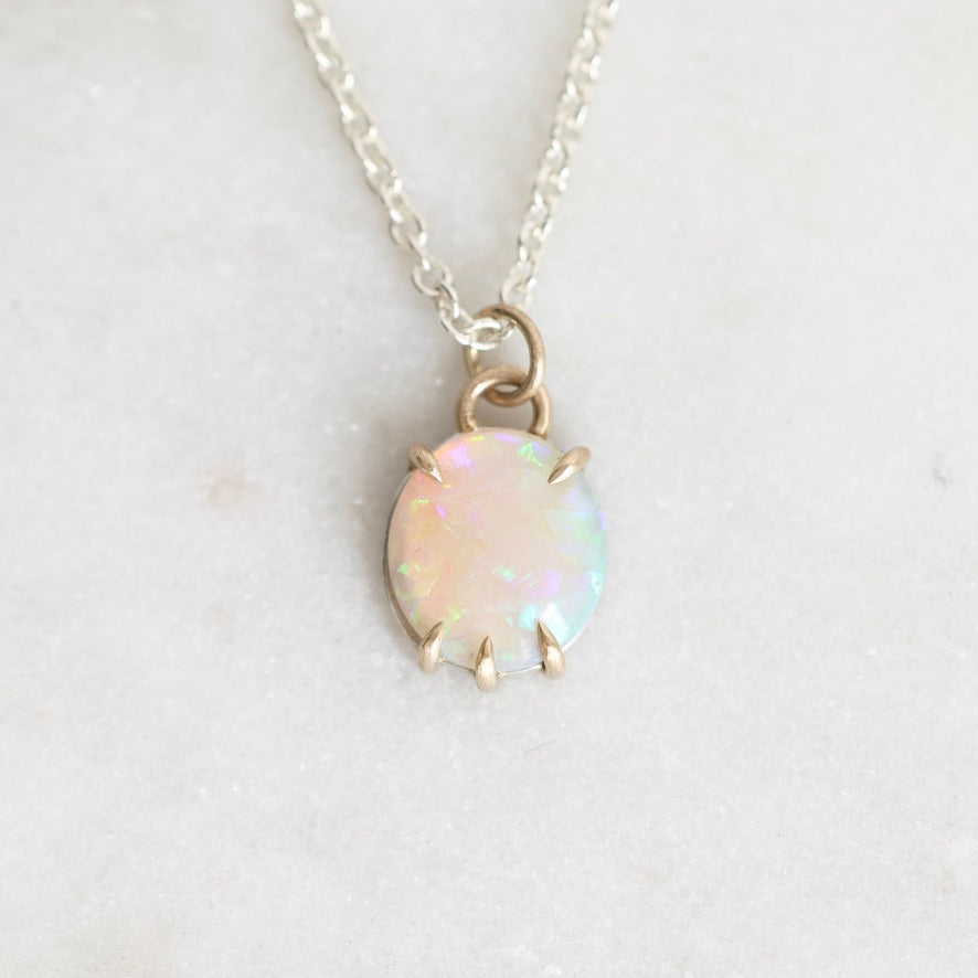 A delicate silver chain necklace with an oval opal pendant in a gold-toned prong setting, resting on a light gray textured surface.