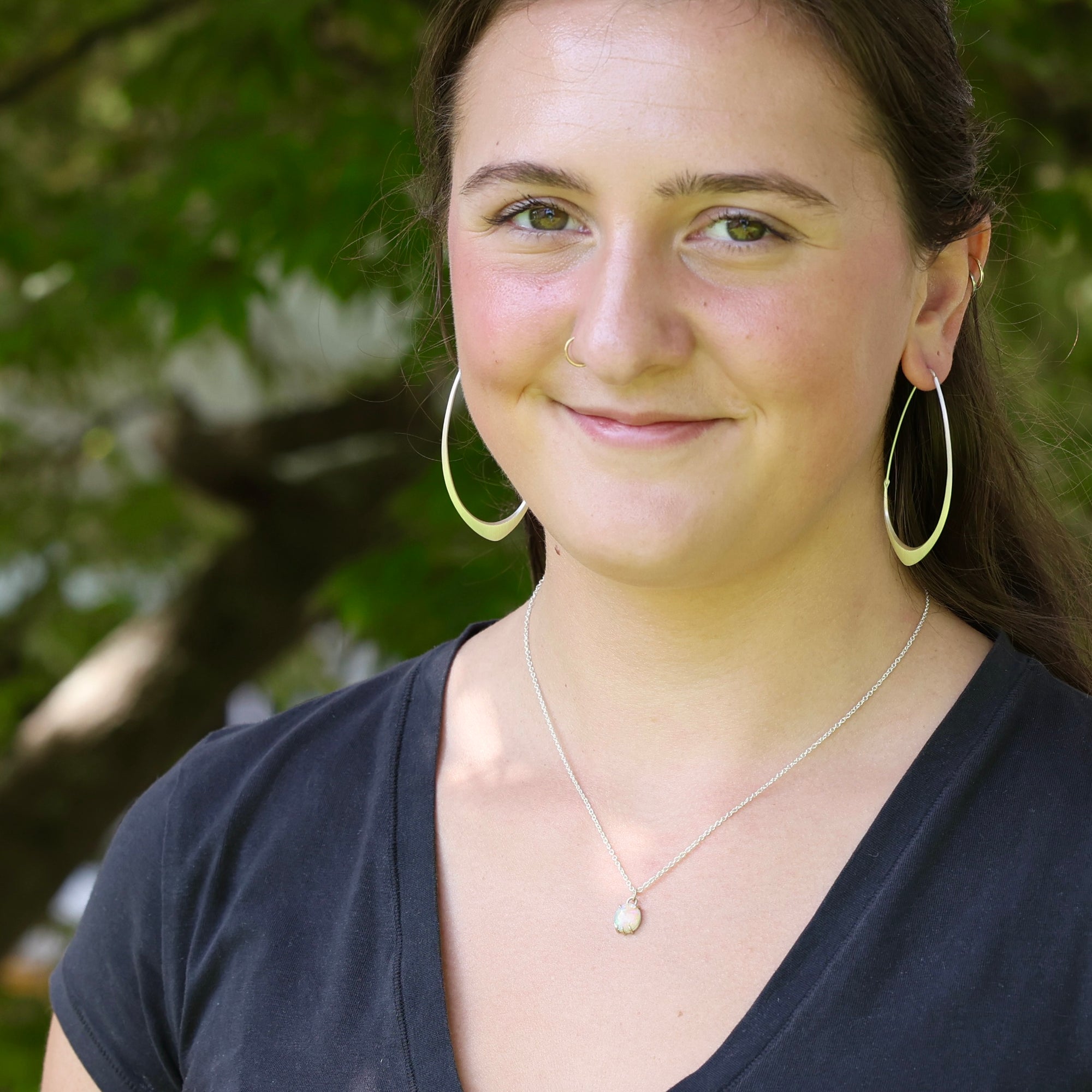 A smiling young woman outdoors wearing large gold hoop earrings, a delicate silver pendant necklace, and a dark navy blue V-neck top, with a soft-focus background of green foliage.