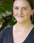 A smiling young woman outdoors wearing large gold hoop earrings, a delicate silver pendant necklace, and a dark navy blue V-neck top, with a soft-focus background of green foliage.