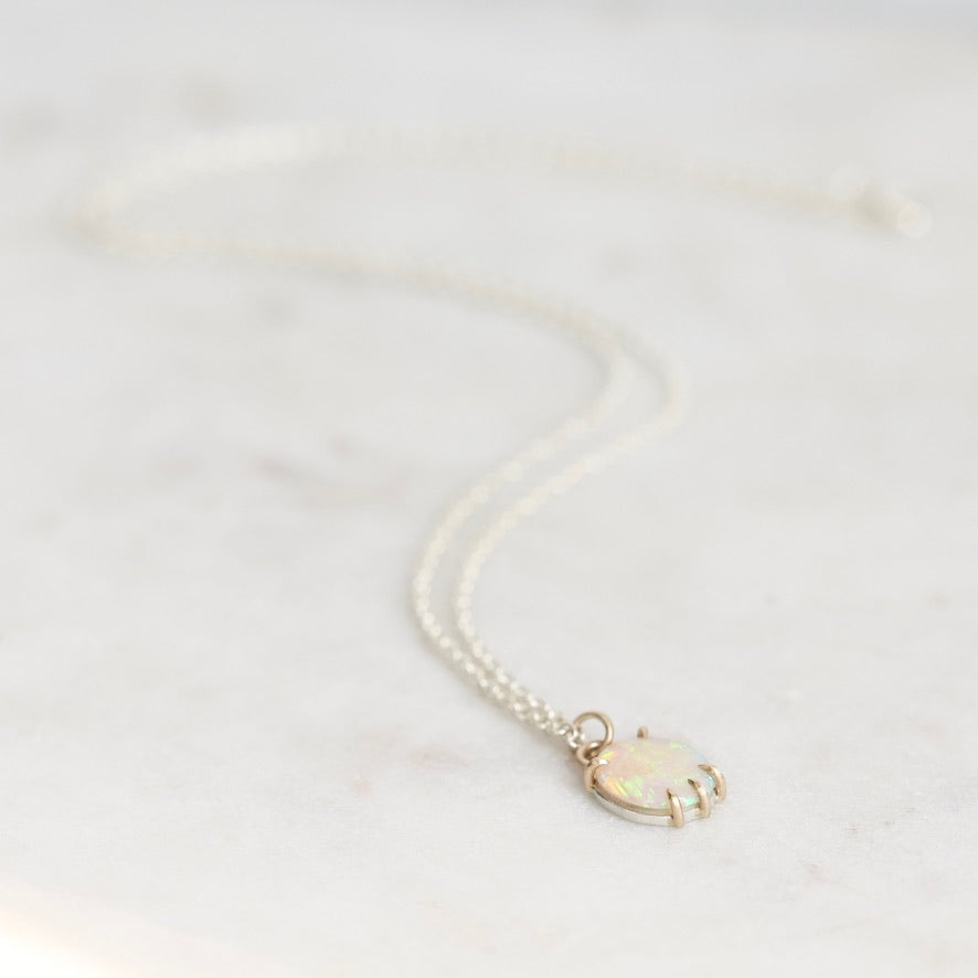 A delicate double-strand necklace with a small oval pendant featuring an iridescent white opal and gold-toned metal, resting on a light marble surface.